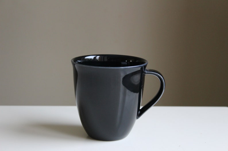 Puede incluir: Una taza de cer&aacute;mica negra con forma redondeada y asa.
