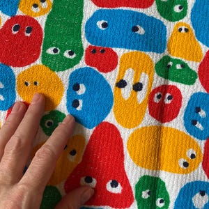 Pu&ograve; includere: Un tessuto bianco con un motivo ripetuto di forme astratte colorate che assomigliano a facce di cartoni animati. Le forme sono rosse, gialle, blu e verdi, ognuna con dettagli di occhi neri e bianchi. Il tessuto ha un aspetto strutturato.