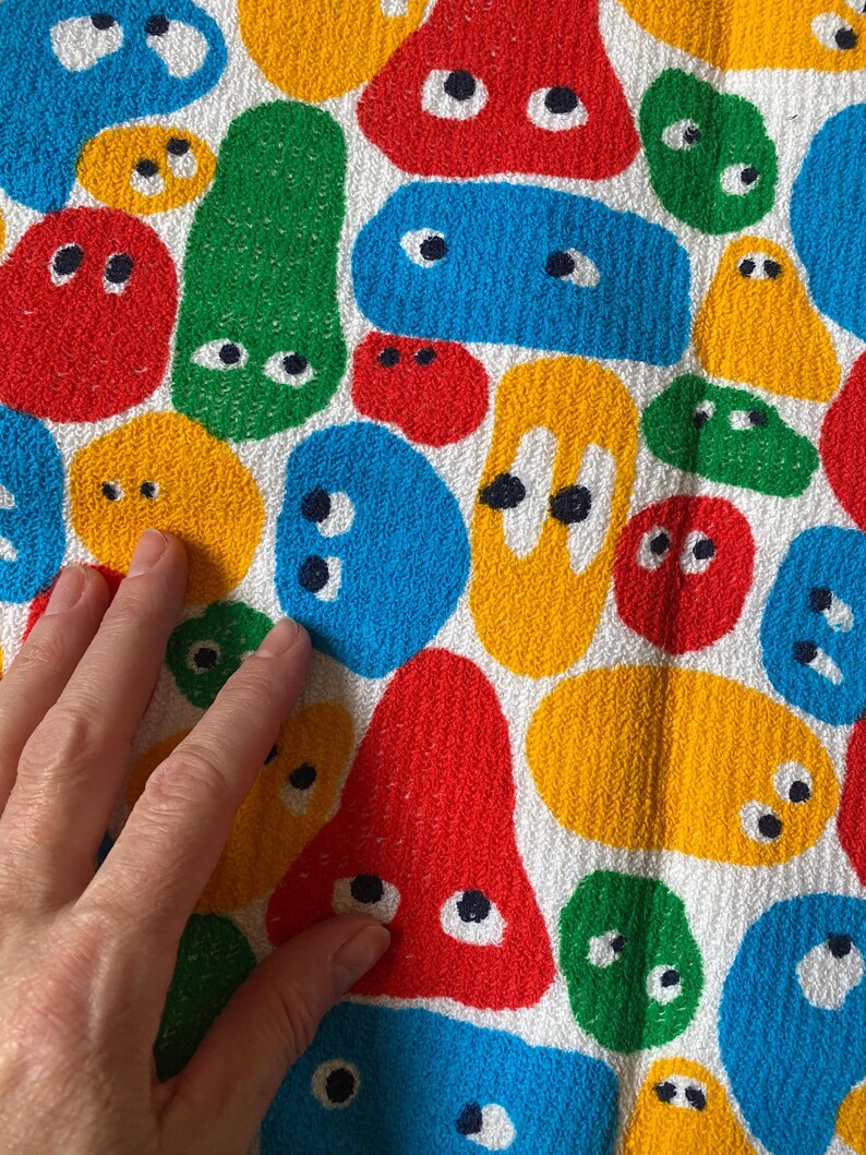 Pu&ograve; includere: Un tessuto bianco con un motivo ripetuto di forme astratte colorate che assomigliano a facce di cartoni animati. Le forme sono rosse, gialle, blu e verdi, ognuna con dettagli di occhi neri e bianchi. Il tessuto ha un aspetto strutturato.