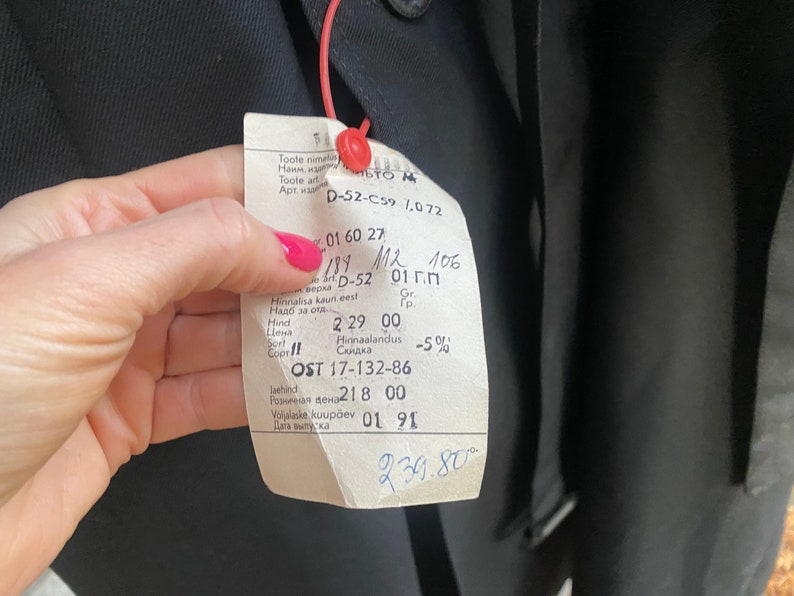Op de afbeelding: Een wit label met zwarte tekst bevestigd aan een zwart kledingstuk. Het label bevat productinformatie zoals artikelnummer, maat, prijs en productiedatum. De tekst op het label luidt: "Toote nimetus", "Toote arti", "Art. на 0-52-C59 1.0 72", "01 60 27", "189 M2 106", "Верка D-52 01 Г.П", "Hinnalisa kaun eest", "Надб за отд", "Hind 2 29 00", "Цена", "Sort 11", "Сорт", "Hinnaalandus -50%", "OST 17-132-86", "laehind", "Нива дена 218 00", "Vollnie kuup&auml;ev 01 91", "Дата выпуска", "239.80".