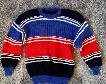 Op de afbeelding: Een gestreepte trui in blauw, rood, wit en zwart. De trui heeft een ronde hals en lange mouwen.