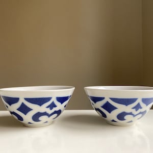 May include: Two white ceramic bowls with a blue and white geometric pattern. The bowls are empty and sitting on a white surface.