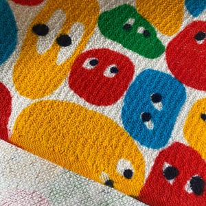 Pu&ograve; includere: Primo piano di un asciugamano in spugna bianco con forme astratte colorate. Le forme sono rosse, gialle, blu e verdi, ciascuna con occhi a puntini neri. L'asciugamano ha una superficie strutturata e un bordo piegato.