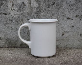 May include: A white ceramic mug with a handle. The mug is empty and sitting on a gray concrete surface.