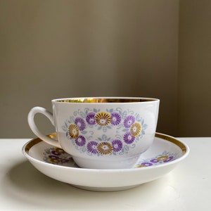 May include: A white teacup and saucer set with a gold rim and a floral pattern of purple and yellow flowers.
