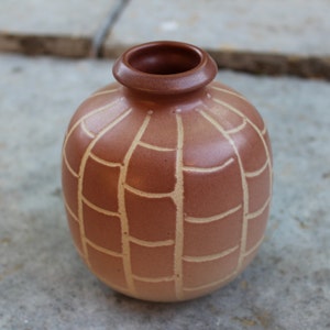Könnte beinhalten: Kleine, runde, braune Keramikvase mit einem strukturierten, gitterartigen Muster in einem helleren Beigeton. Die Vase hat einen schmalen Hals und einen leicht ausgestellten Rand, geeignet für kleine Blumen oder Dekorationsartikel.