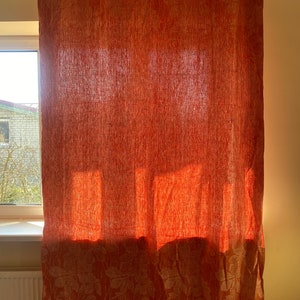 May include: Orange linen curtains with a white floral pattern. The curtains are hanging in a window and the sunlight is shining through them.