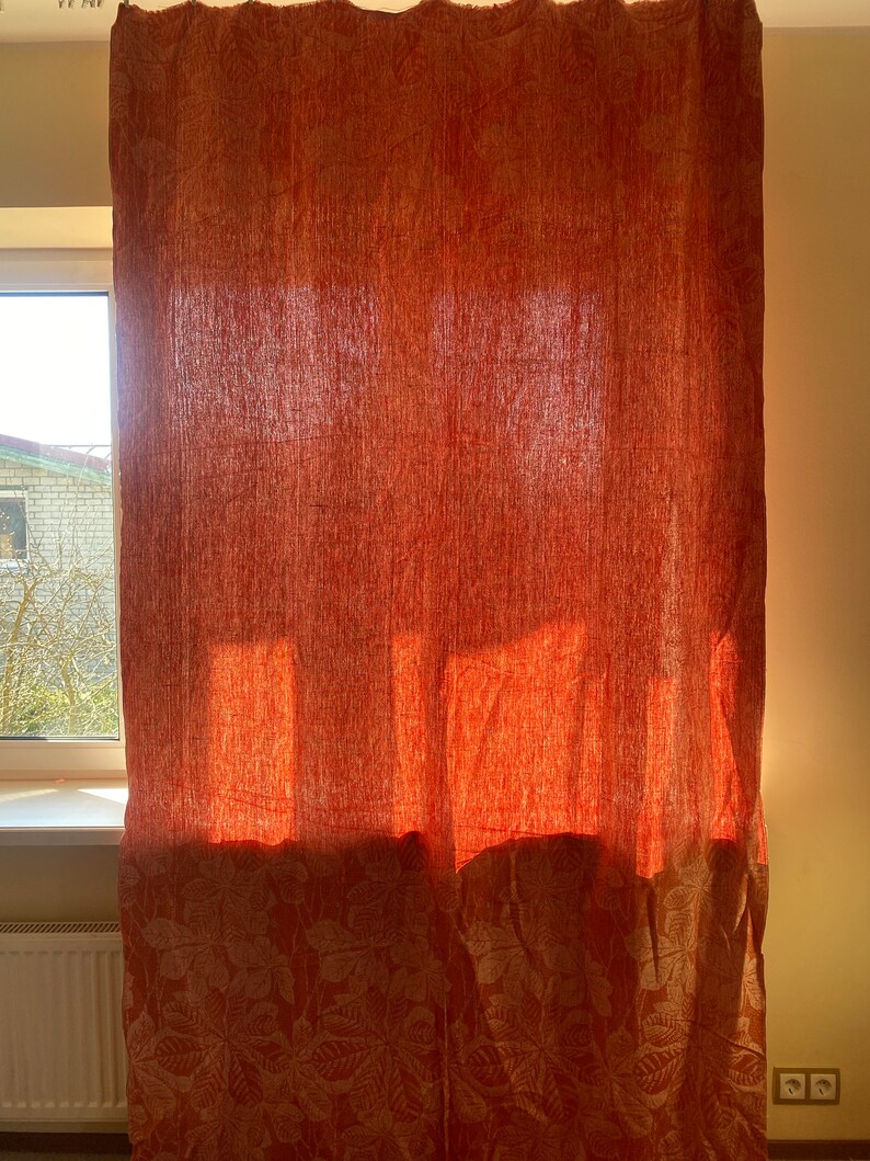 May include: Orange linen curtains with a white floral pattern. The curtains are hanging in a window and the sunlight is shining through them.