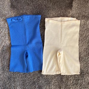 May include: Two pairs of vintage ribbed knit underwear. One pair is a bright blue, and the other is a cream color. Both pairs have a high waist and short legs. The blue pair has a bow detail at the waist.