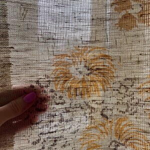 May include: A close-up of a beige sheer curtain with a woven texture and a brown floral pattern. The curtain has a subtle sheen and a delicate, airy feel.