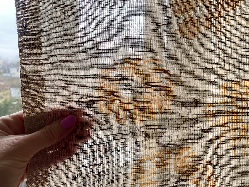 May include: A close-up of a beige sheer curtain with a woven texture and a brown floral pattern. The curtain has a subtle sheen and a delicate, airy feel.
