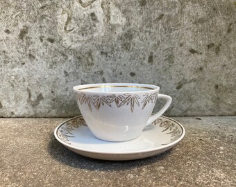 May include: A white teacup and saucer with a gold leaf design. The cup has a handle and the saucer is round.