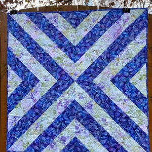 May include: A quilt with a geometric pattern of diagonal stripes in shades of blue and white. The fabric has a floral print with a subtle purple hue.