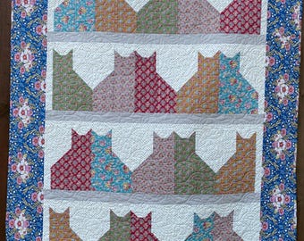 May include: A quilt with a blue floral border and a white background featuring a pattern of colorful cats. The cats are arranged in rows of three, with each cat made up of five different fabric squares.