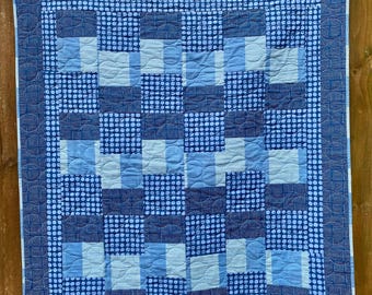 May include: A handmade quilt in shades of blue, featuring a patchwork design of squares and dotted patterns. The quilt is displayed outdoors, hanging on a wooden fence. The quilt's dimensions are not visible.