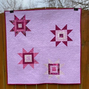 May include: A purple quilt with four pink and white star-shaped blocks. The quilt is hanging on a wooden fence.