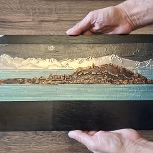 Peut inclure: Sculpture en relief rectangulaire représentant un paysage avec des montagnes, de l'eau et une ville. L'œuvre a un cadre noir et est tenue par deux mains. Les montagnes sont argentées, l'eau est turquoise et la ville est bronze.