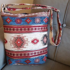 May include: A rectangular shoulder bag with a colorful woven pattern. The bag features a cream-colored central panel with a geometric design in red, pink, and gray. Blue and red bands with geometric shapes border the central panel. The strap is cream-colored with red accents.