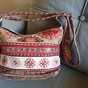 May include: A shoulder bag with a woven fabric design in shades of red, beige, and brown. The bag features a repeating geometric pattern and a long strap. The interior is black.