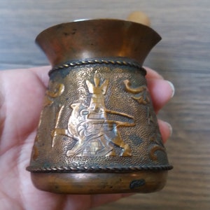 May include: A small, antique copper-colored coffee grinder with a wooden handle. The grinder features an embossed design of a Native American figure and decorative scrollwork. Braided trim accents the top and bottom.