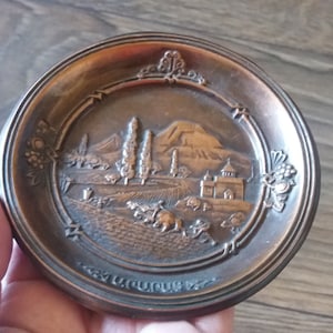 Puede incluir: Un plato decorativo pequeño y redondo de color bronce con una escena en relieve detallada de un paisaje con edificios, árboles y montañas. El plato tiene un borde decorativo con racimos de uvas y texto en la parte inferior.