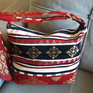 May include: A patterned shoulder bag with a red strap and handle. The bag features a mix of red, black, white, and gold geometric designs. The bag is sitting on a gray surface.