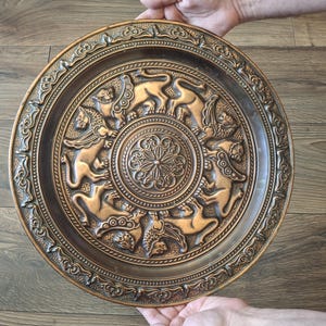 May include: A round, copper-colored decorative plate with an embossed design. The plate features a central floral pattern surrounded by a ring of stylized animal figures. The outer rim has a detailed, repeating pattern. The plate is held by two hands.