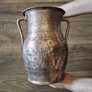 May include: A large, hand-hammered copper pitcher featuring two side handles and an intricate floral band engraved around the neck. The body displays a mottled, aged patina with horizontal lines, showcasing a wide mouth and a tapered base.