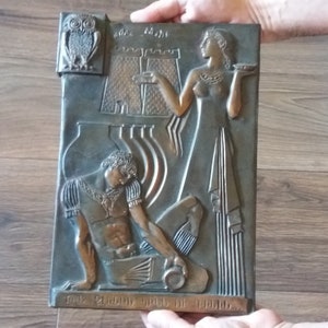 May include: Bronze relief plaque featuring classical figures and architectural elements. An owl is depicted in the upper left corner. The plaque shows a woman holding a plate and a man reclining, with text at the bottom.