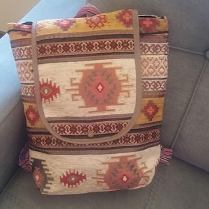 May include: A patterned backpack with a flap closure. The bag features a beige background with geometric designs in red, brown, and yellow. The straps and trim are a solid brown color. The backpack is sitting on a gray surface.