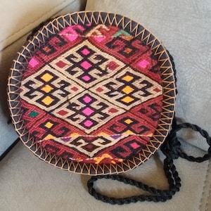 May include: A round, woven bag with a black strap. The bag features a geometric pattern in red, pink, yellow, and black. The edges are stitched with a contrasting thread. The bag is approximately 8 inches in diameter.