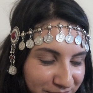 Puede incluir: Diadema plateada con colgantes de monedas. La diadema presenta una piedra roja central y una serie de pequeñas monedas redondas con un perfil. Un accesorio decorativo para el cabello.