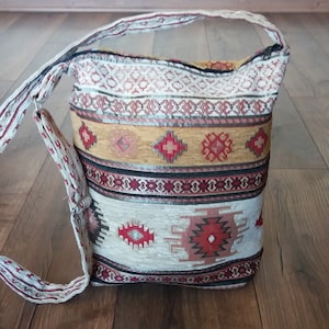 May include: A rectangular shoulder bag with a patterned design in cream, red, black, and gold. The bag has a long strap and a matching patterned design. The bag is made of a woven fabric.