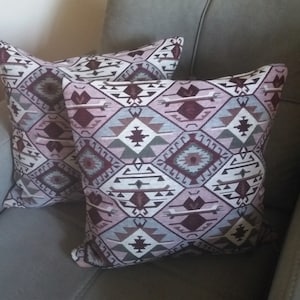 May include: Two square throw pillows featuring a repeating geometric diamond pattern in shades of dusty rose, deep burgundy, slate blue, and cream. The woven fabric displays intricate symmetrical shapes and lines across the entire surface.