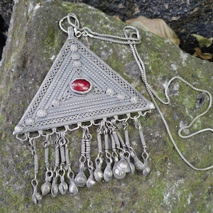 Puede incluir: Un colgante triangular de plata con intrincados trabajos de filigrana y una piedra roja ovalada en el centro. El colgante cuelga de una fina cadena de plata e incluye múltiples dijes de plata colgantes a lo largo del borde inferior.