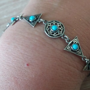 Puede incluir: Una pulsera de metal plateado que presenta una serie de formas geométricas alternas, incluyendo triángulos y un medallón circular central, cada uno incrustado con una pequeña piedra turquesa redonda y brillante. El metal tiene un acabado oxidado.