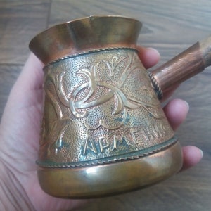 May include: A copper cezve, a traditional coffee pot, with ornate floral engravings and the word "ARMENIA" etched on the side. It features a wooden handle and a detailed, textured design. The pot has a vintage, antique appearance.