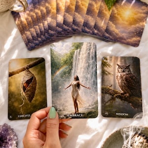 May include: A deck of oracle cards with a celestial sky design, featuring three face-up cards: one showing a chrysalis labeled UNRAVEL, one showing a person standing before a waterfall labeled EMBRACE, and one showing an owl labeled WISDOM.