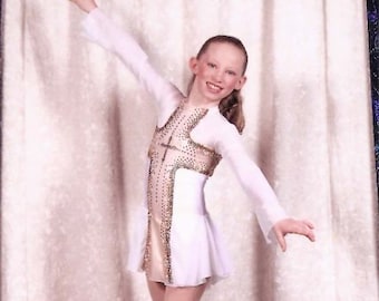 May include: A young figure skater in a white and gold skating dress with a cross design. She is posing on the ice with her right leg extended and her left leg bent. The background is a white curtain with a blue border.
