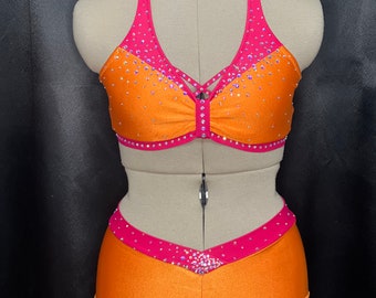 May include: Orange and pink dance costume with rhinestones. The top is a halter style with a criss-cross design in the front. The bottom is a pair of shorts with a V-shaped design in the front.
