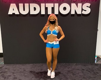 Könnte beinhalten: Eine Frau in einem blauen und weißen Tanzkostüm mit einer weißen Gesichtsmaske steht vor einer schwarzen Wand mit pinkfarbenen Neonbuchstaben, auf denen "Football's Fabulous Females AUDITIONS" steht.