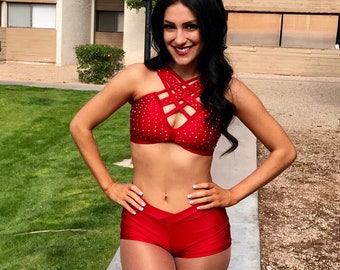 May include: A woman wearing a red, sequined, halter-style top with a criss-cross design and matching red shorts. She is standing on a sidewalk with green grass in the background.
