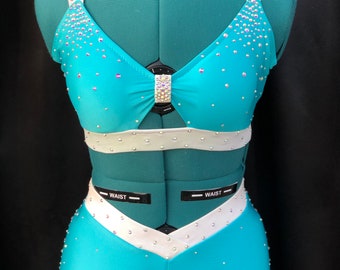 May include: A turquoise dance costume with a white trim. The top has a bow in the center and the shorts have a V-shaped white trim. The costume is decorated with rhinestones.