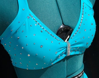 Könnte beinhalten: Ein türkisfarbenes Tanzkostüm mit einem glitzernden Oberteil und passenden Hosen. Das Oberteil hat einen herzförmigen Ausschnitt und ein Schleifendetail in der Mitte. Die Hose hat eine hohe Taille und ist mit Strasssteinen verziert.