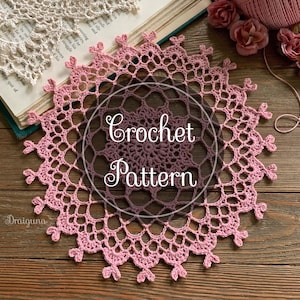 Op de afbeelding: Een roze gehaakt tafeltje met een delicaat patroon. Het tafeltje ligt op een houten oppervlak en de tekst "Crochet Pattern" is over de afbeelding gelegd.