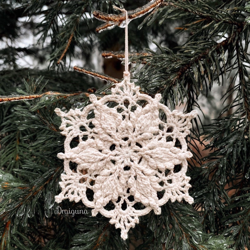 May include: A white crocheted snowflake ornament hanging from a branch of a pine tree covered in ice.