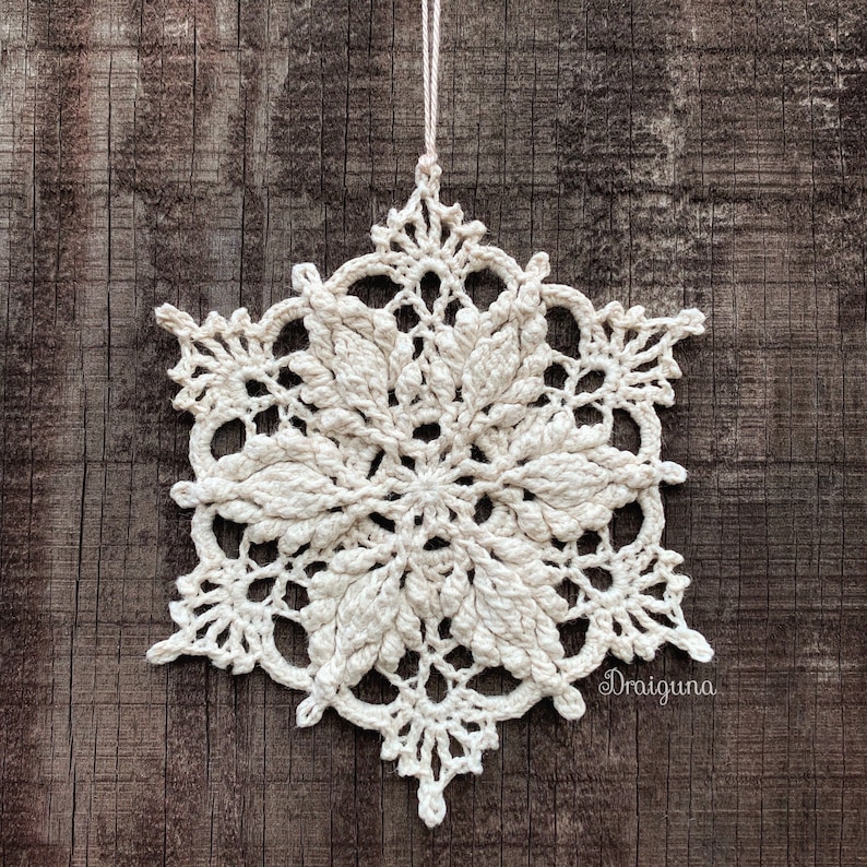 May include: A white crocheted snowflake ornament hanging from a string against a brown wooden background. The snowflake has a delicate, intricate design.