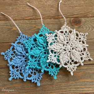 May include: Three crocheted snowflakes in shades of blue and white. The snowflakes are hanging from white strings and are arranged in a row on a wooden surface.