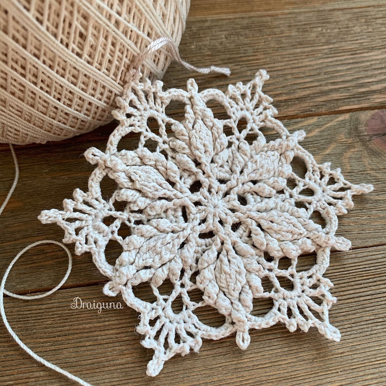 May include: A delicate crocheted snowflake in a cream color, with intricate details and a lacy design. The snowflake is lying on a wooden surface.
