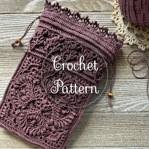 Op de afbeelding: Een gehaakte trekkoordtas in een diepe mauve kleur, met ingewikkelde bloempatronen. De tas heeft een decoratieve geschulpte rand en een trekkoord sluiting met houten kralen. De tekst "Crochet Pattern" is zichtbaar.